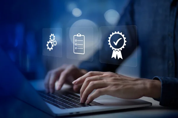 Manos en un portátil con iconos de engranajes, una lista de comprobación y un sello de certificación